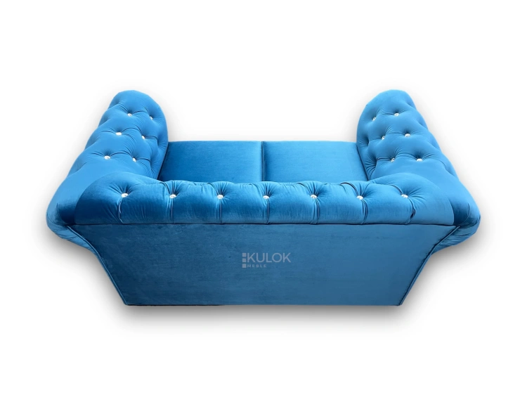 Elegancka niebieska sofa dwuosobowa z pikowanym oparciem w stylu Chesterfield, tapicerowana miękkim welurem, ozdobiona dekoracyjnymi guzikami.